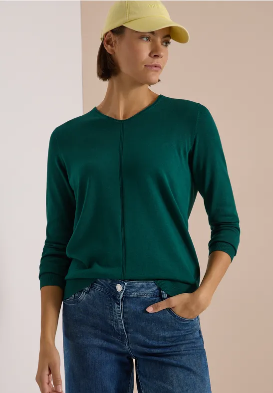 Frau mit gelber Mütze und grünem Pullover, blaue Jeans, seitlich stehend vor hellem Hintergrund.