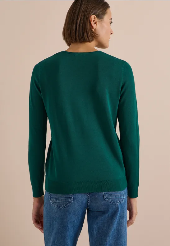 Rückansicht einer Frau in grünem Pullover und blauen Jeans vor beigem Hintergrund.