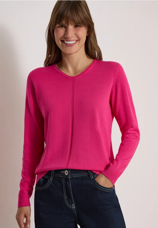 Lächelnde Frau im pinken V-Pullover und dunklen Jeans, Hände in den Taschen.
