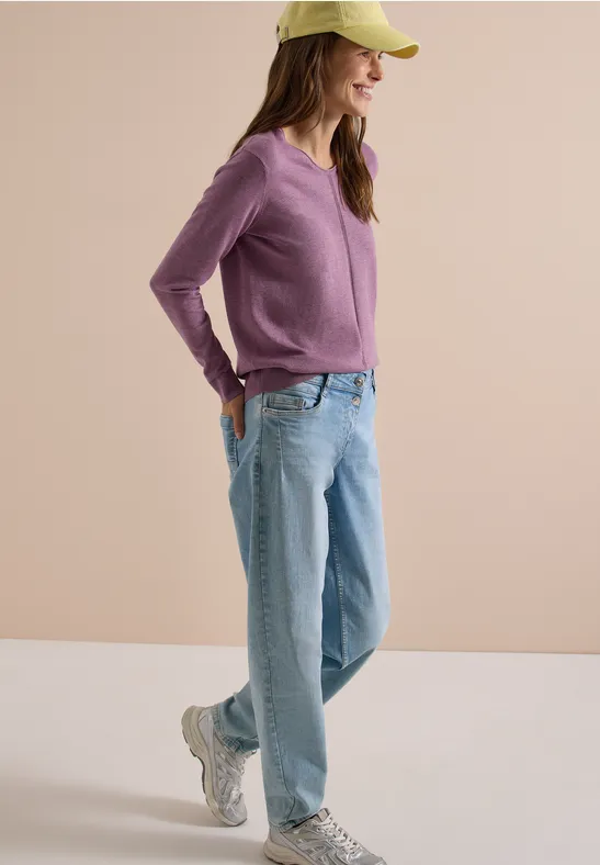 Eine Frau mit grüner Cap, lila Pullover, hellblauen Jeans und Sneakern lächelt entspannt mit Händen in den Taschen.