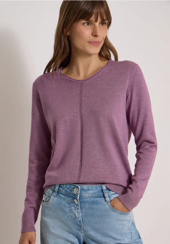 Junge Frau im violetten Pullover mit vertikalem Ziernaht und Jeans, Hände in Hosentaschen.