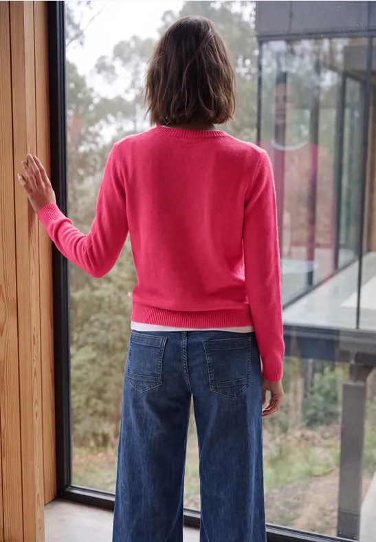 Rückansicht einer Person in pinkem Pullover und blauer Jeans vor einem Fenster mit Waldblick.