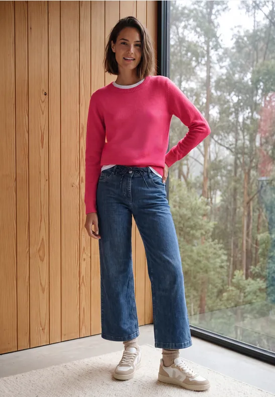 Frau in pinkem Pullover und Jeans vor Holzwand und Fenster mit Waldblick.