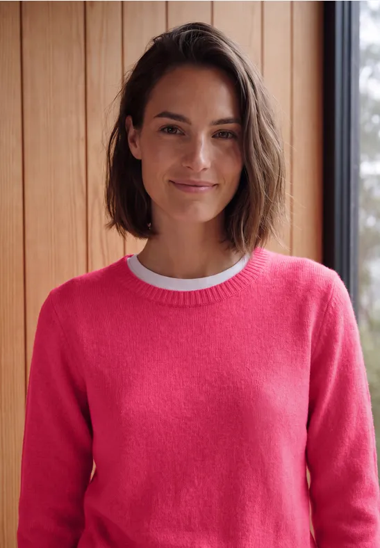 Frau mit dunklen Haaren lächelt und trägt einen pinken Pullover vor Holzwand.
