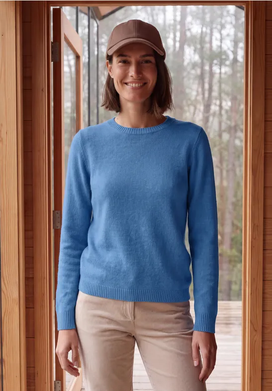 Lachende vrouw in blauw truitje, beige broek en pet, staand voor een houten raam.