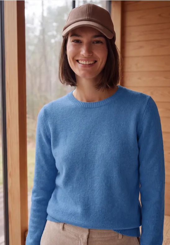 Jonge vrouw met blauwe trui en beige pet voor een houten wand, ze lacht.