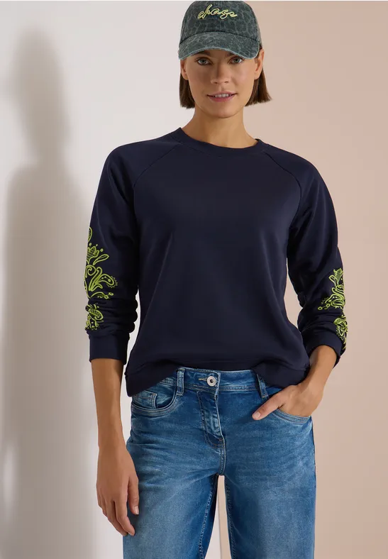 Frau in navyblauem Sweatshirt mit neongrünen Blumenstickereien an den Ärmeln, Jeans und Basecap.