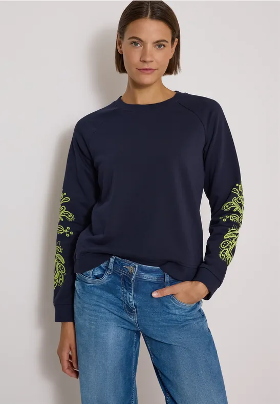 Frau in dunkelblauem Sweatshirt mit grünen Ärmelmustern und Jeans vor hellem Hintergrund.