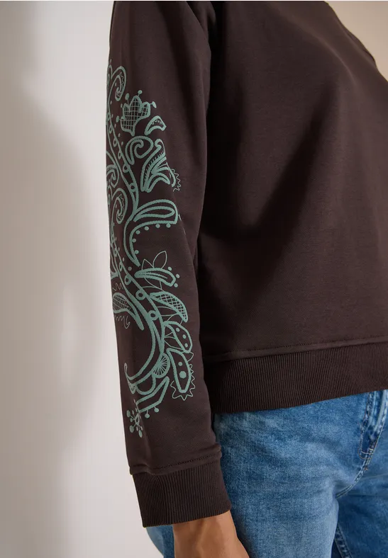 Schwarzer Pullover mit türkisem Paisleymuster am Ärmel und blaue Jeans.