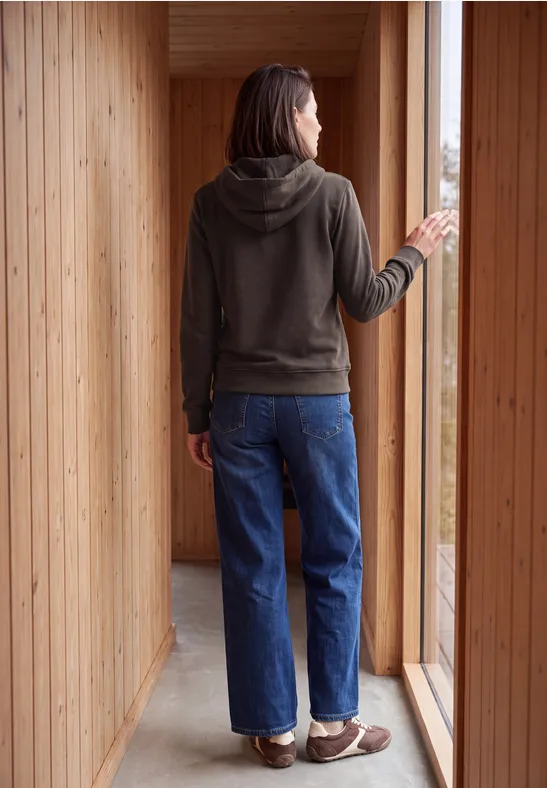 Frau in Hoodie und Jeans, von hinten in einem Holzraum stehend, blickt aus einem großen Fenster.