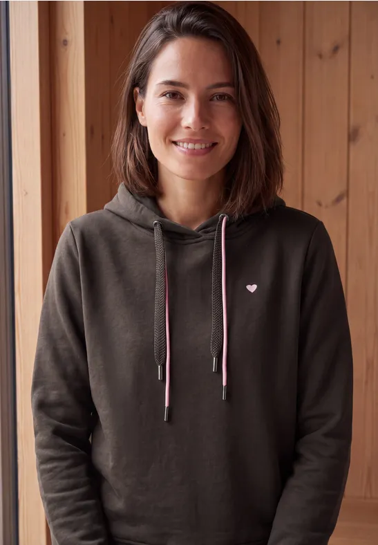 Eine lächelnde Frau in dunkelgrauer Kapuzenjacke mit kleinem Herzlogo vor Holzpaneelen.