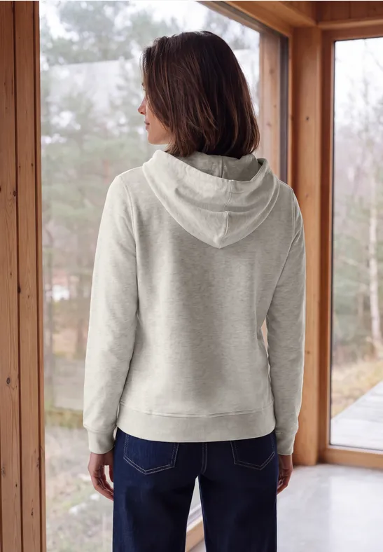 Frau von hinten in grauem Kapuzenpullover und Jeans, stehend vor großen Fenstern mit Blick ins Freie.