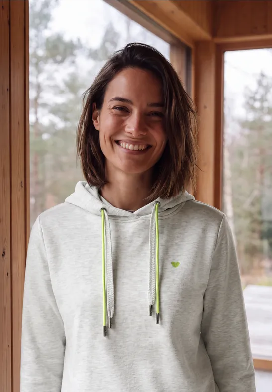 Eine lächelnde Frau in grauem Kapuzenpullover mit grünen Kordeln steht an einem Fenster in Holzhütte.