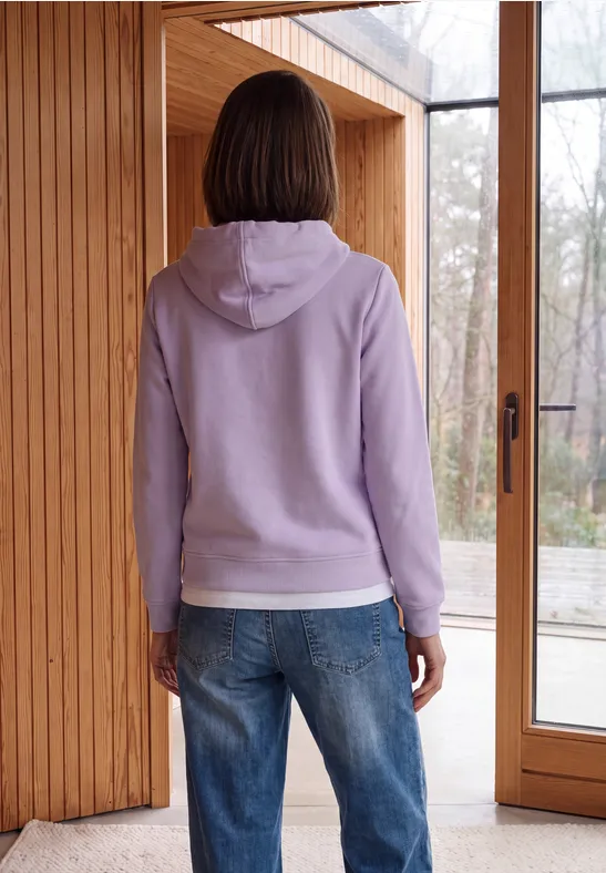 Eine Person in lila Kapuzenpullover und Jeans steht vor einer großen Glasschiebetür mit Holzrahmen.