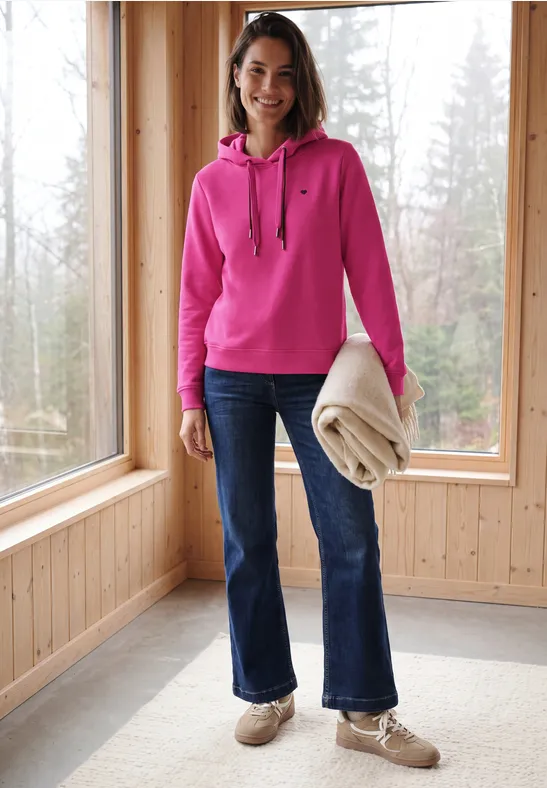 Frau in pinkem Hoodie, Jeans und beigen Schuhen hält Decke in einem hellen Holzraum mit großen Fenstern.
