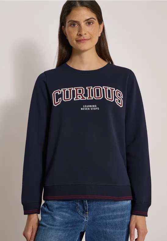 Junge Frau trägt dunkelblaue Sweatshirt mit Aufschrift 'CURIOUS', lächelt vor neutralem Hintergrund.