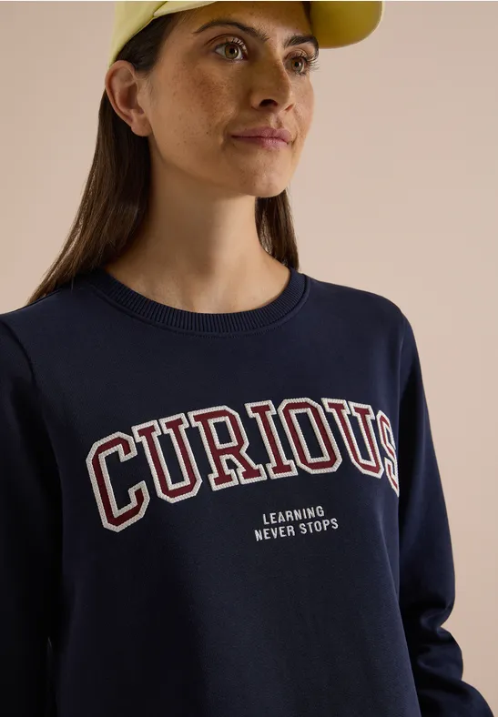 Eine junge Frau mit gelber Mütze und dunkelblauem Sweatshirt mit Aufschrift 'CURIOUS' blickt seitlich.