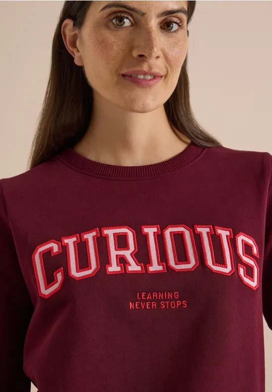 Frau in bordeauxrotem Pullover mit großem „CURIOUS“-Schriftzug und kleinem „Learning Never Stops“-Motto lächelnd