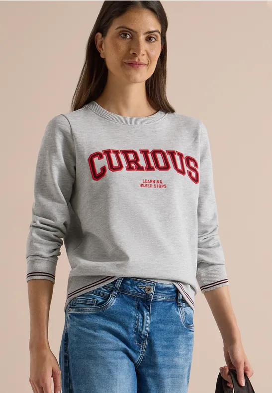 Junge Frau in grauem Sweatshirt mit rotem 'CURIOUS'-Schriftzug und blauen Jeans.
