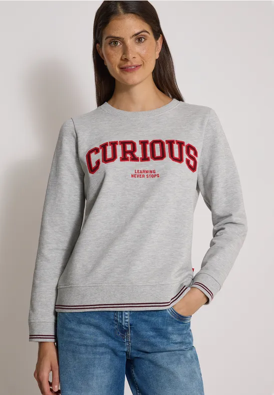 Eine Frau trägt ein graues Sweatshirt mit rotem Schriftzug 'CURIOUS' und blaue Jeans.