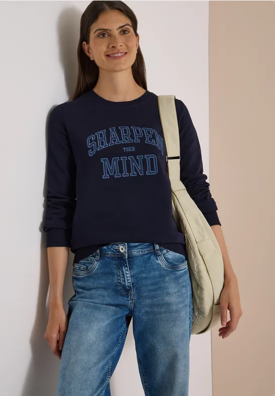 Frau in Jeans und dunkelblauem Pullover mit Aufschrift 'SHARPEN MIND', trägt eine Tasche und lächelt