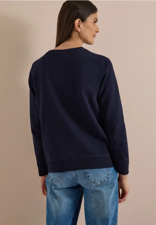 Rückansicht einer Frau in dunkelblauem Pullover und Jeans vor beigem Hintergrund.
