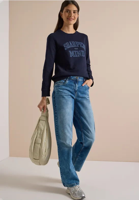 Frau in lässigem Outfit: dunkelblauer Pullover mit Aufschrift, Jeans, Sneaker und helle Schultertasche.