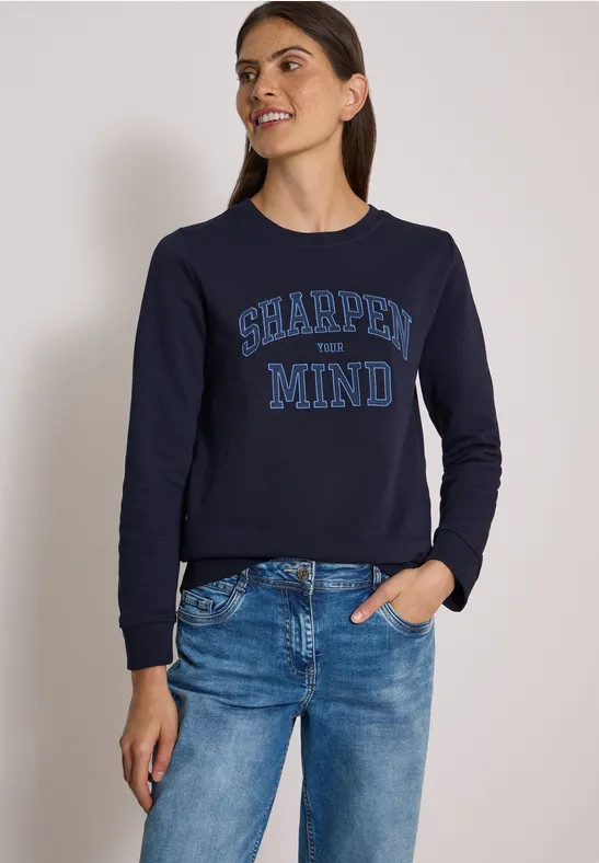 Frau trägt dunkelblauen Pullover mit 'SHARPEN MY MIND'-Schriftzug und Jeans vor neutralem Hintergrund.
