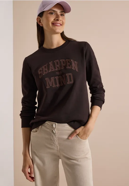 Frau in dunkelgrauem Sweatshirt mit 'SHARPEN MIND'-Aufdruck, beigen Jeans und rosa Mütze lächelt.