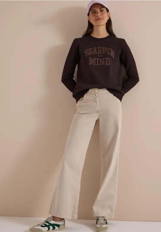 Frau trägt braunen Pullover mit Aufdruck, beige weite Hose, grüne Sneaker und rosa Cap vor neutralem Hintergrund.