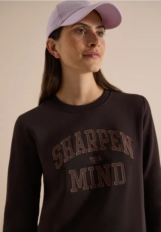Eine Frau trägt eine helle Kappe und einen schwarzen Pullover mit der Aufschrift "SHARPEN MIND".
