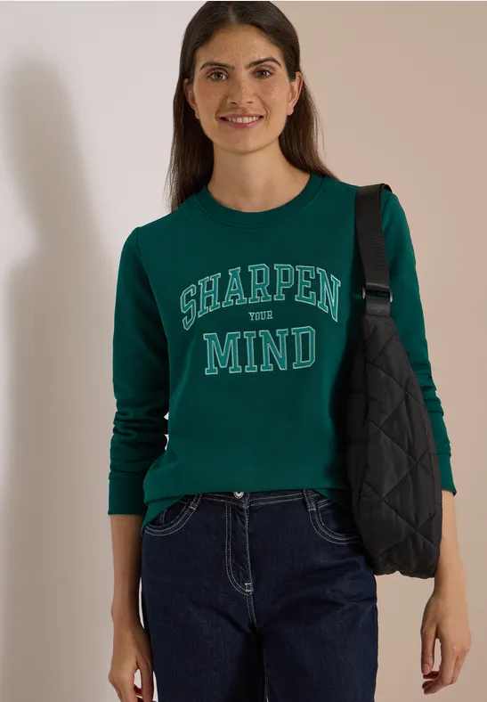 Eine lächelnde Frau trägt einen dunkelgrünen Pullover mit der Aufschrift 'SHARPEN MIND' und eine dunkle Jeans.