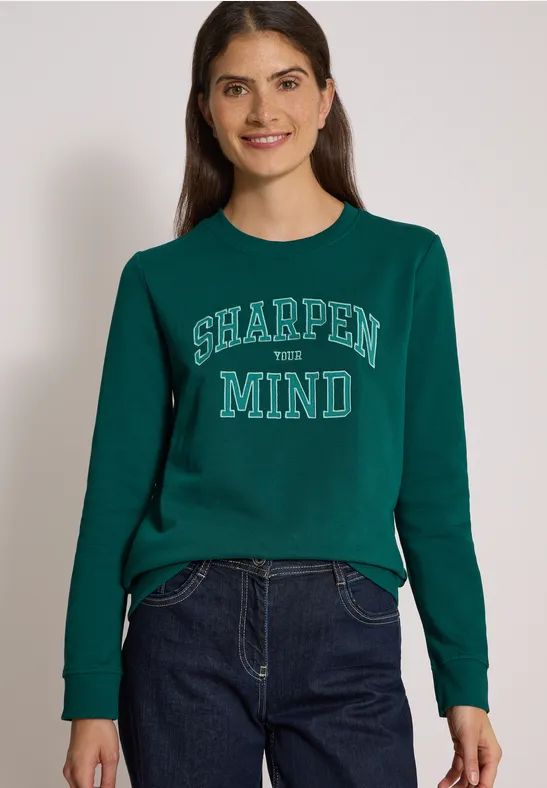 Junge Frau trägt grünes Sweatshirt mit Aufdruck 'SHARPEN YOUR MIND' und blaue Jeans.