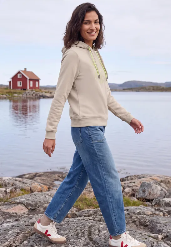 Eine Frau in Hoodie und Jeans spaziert am Seeufer vor einer roten Hütte.