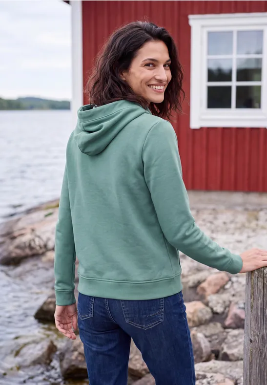 Eine lächelnde Frau in grüner Kapuzenjacke und Jeans steht am Ufer vor einem roten Haus am See.
