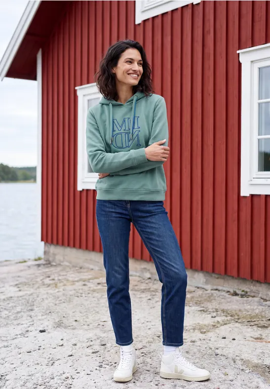 Frau in grüner Kapuzenjacke, Jeans und weißen Stiefeletten steht vor roter Holzhütte am See.
