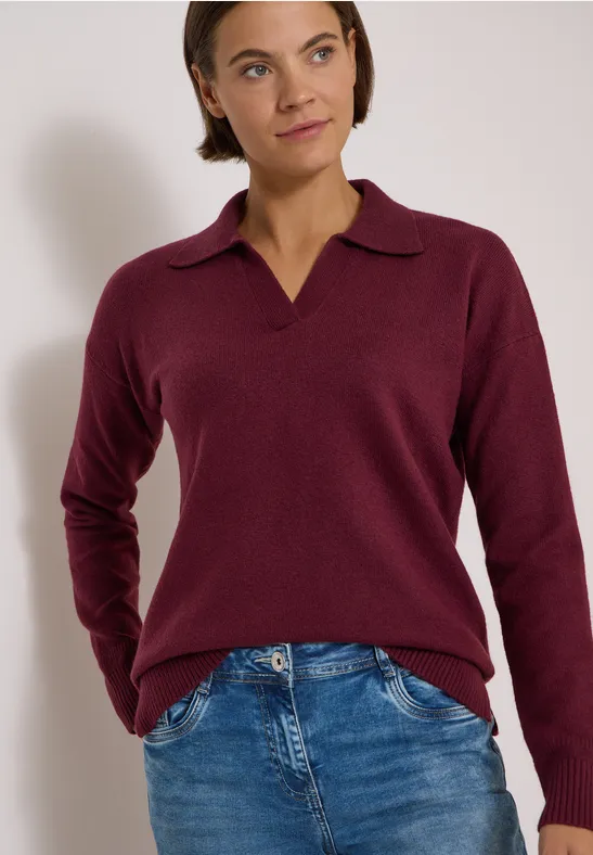 Femme portant un pull bordeaux à col en V et un jean bleu, posant devant un fond blanc.