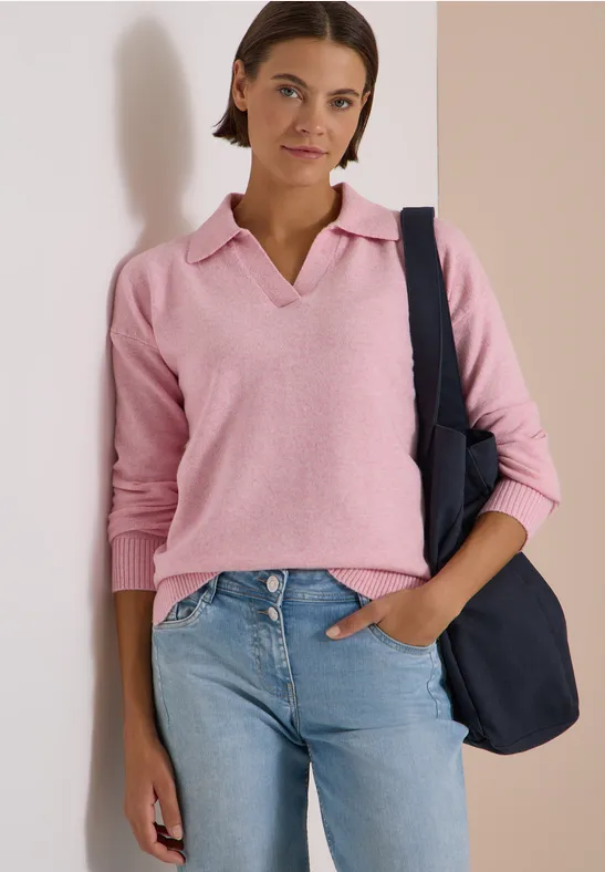 Eine junge Frau trägt ein rosafarbenes Poloshirt, Jeans und eine dunkelblaue Tasche über der Schulter.
