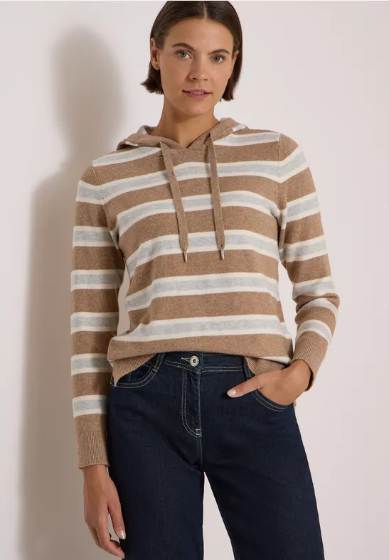 Femme en sweat à capuche rayé beige et blanc avec jean, posant devant un fond neutre.