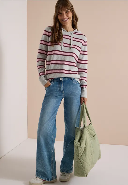 Frau mit gestreiftem Hoodie, weiten Jeans und großer grüner Tasche, lächelnd vor hellem Hintergrund.