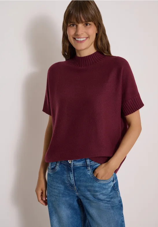 Frau in kurzärmligem, bordeauxrotem Strickpullover und Jeans lächelt vor hellem Hintergrund.