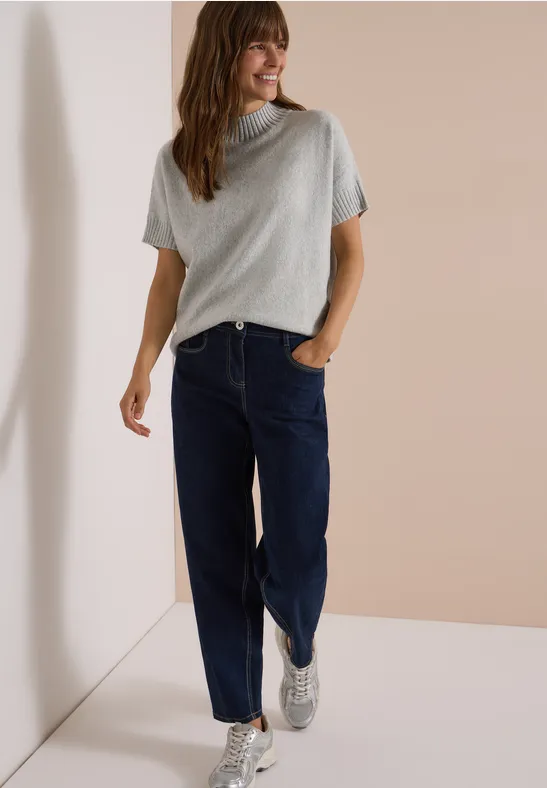 Frau lehnt lässig an Wand und trägt graues T-Shirt, blaue Jeans und weiße Sneaker.