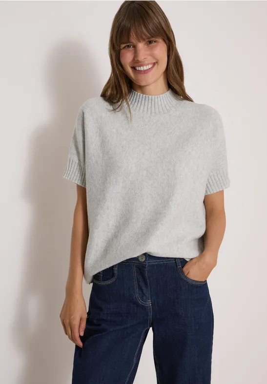 Junge Frau mit braunem Haar, grauem Kurzarm-Pullover und Jeans, lächelnd vor hellem Hintergrund.