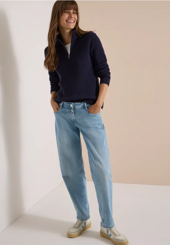 Femme en pull bleu foncé, jean clair et baskets, debout et souriante.