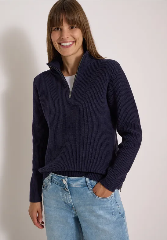 Femme souriante en pull bleu marine et jean, une main dans la poche.