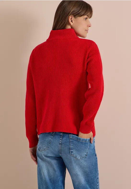 Femme de dos, regard latéral droit, pull col roulé rouge et jean bleu sur fond beige.