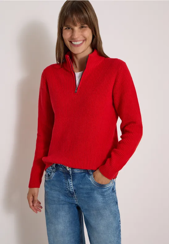Une femme souriante porte un pull rouge et un jean bleu sur fond blanc.