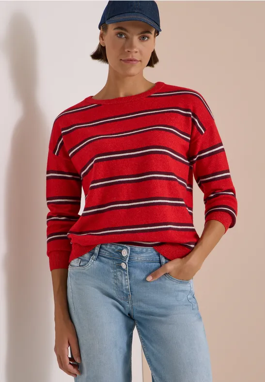Frau mit rotem, schwarz-weiß gestreiftem Pullover, blauer Jeans und Cap vor neutralem Hintergrund.