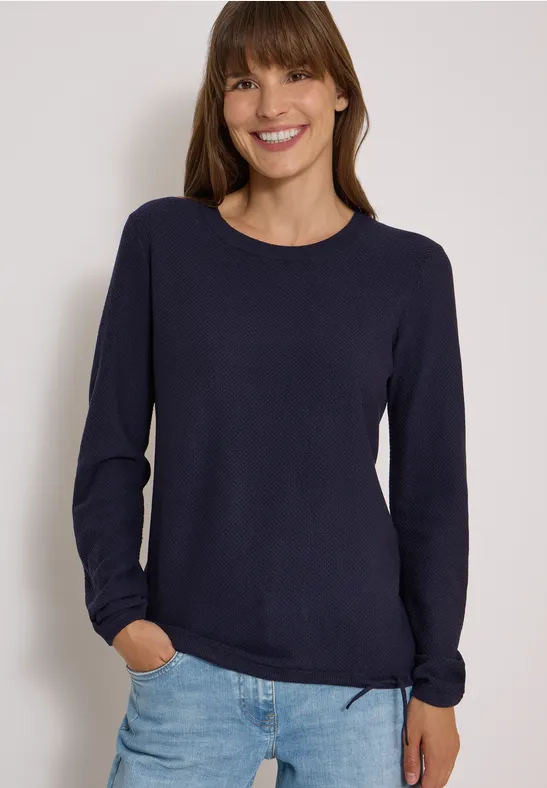 Femme sourit en pull bleu marine et jean clair sur fond blanc.