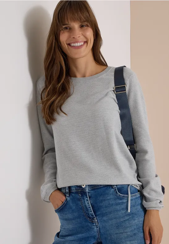 Jeune femme souriante en pull gris, jean bleu et sac à dos, devant un mur bicolore.